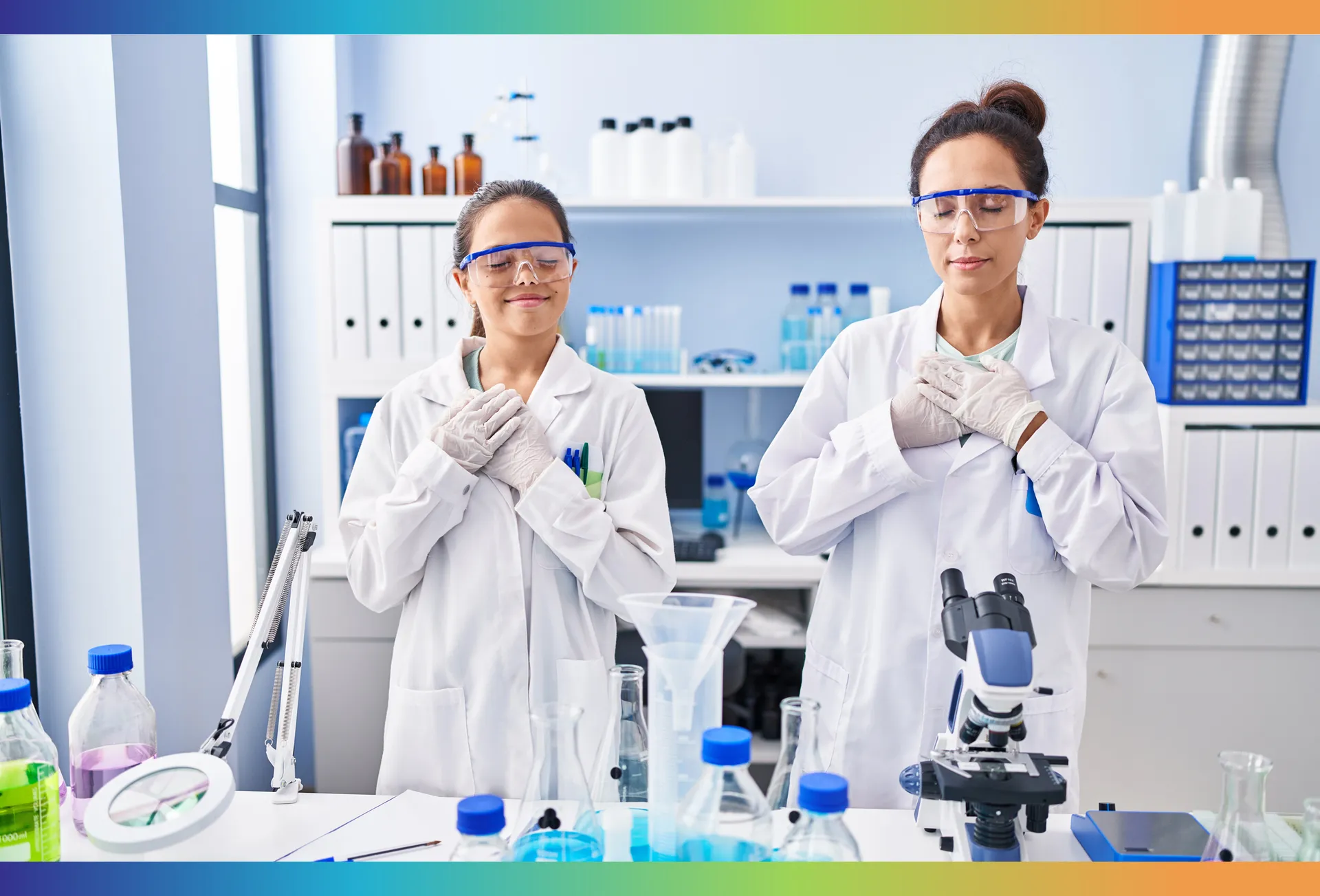 Deux femmes portent des blouses blanches. Elles sont dans un laboratoire. Elles ont les yeux fermés, elles sourient et ont les mains croisées sur leur poitrine.