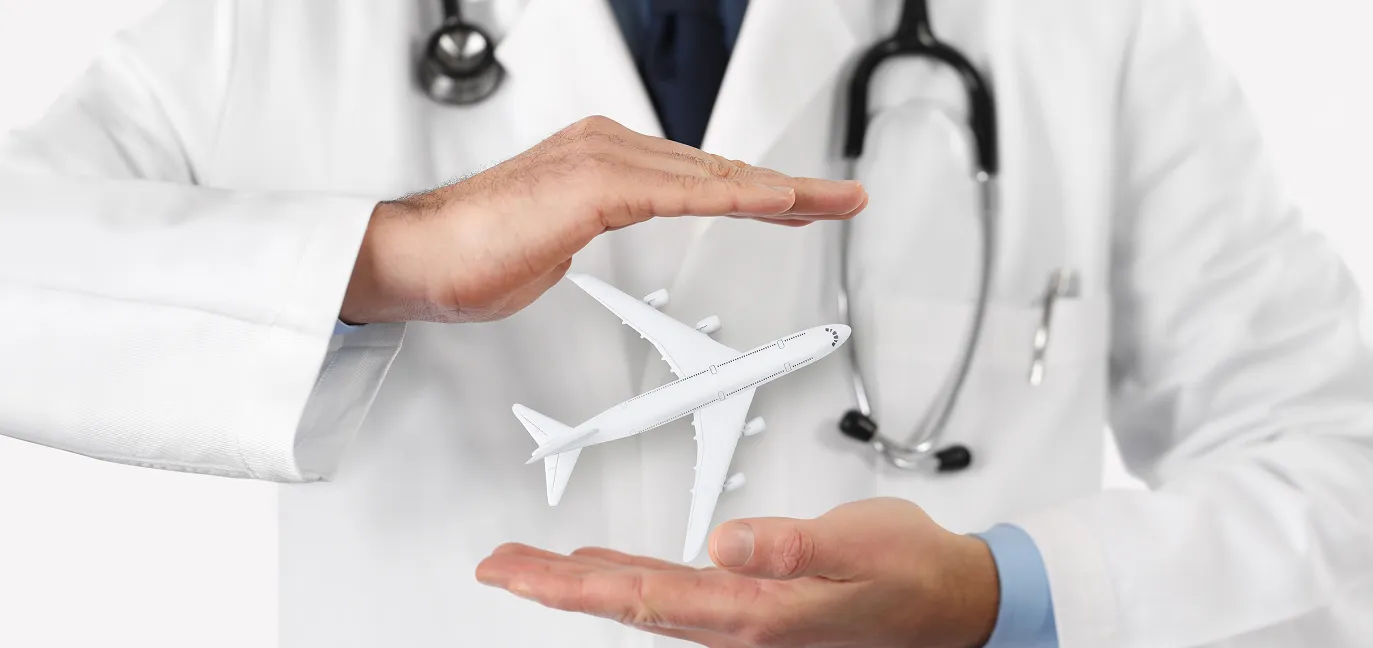 Un médecin porte une blouse blanche et il a un stéthoscope autour de son cou. Ses mains sont l’une au-dessus de l’autre. Entre ses mains, il y a un avion.