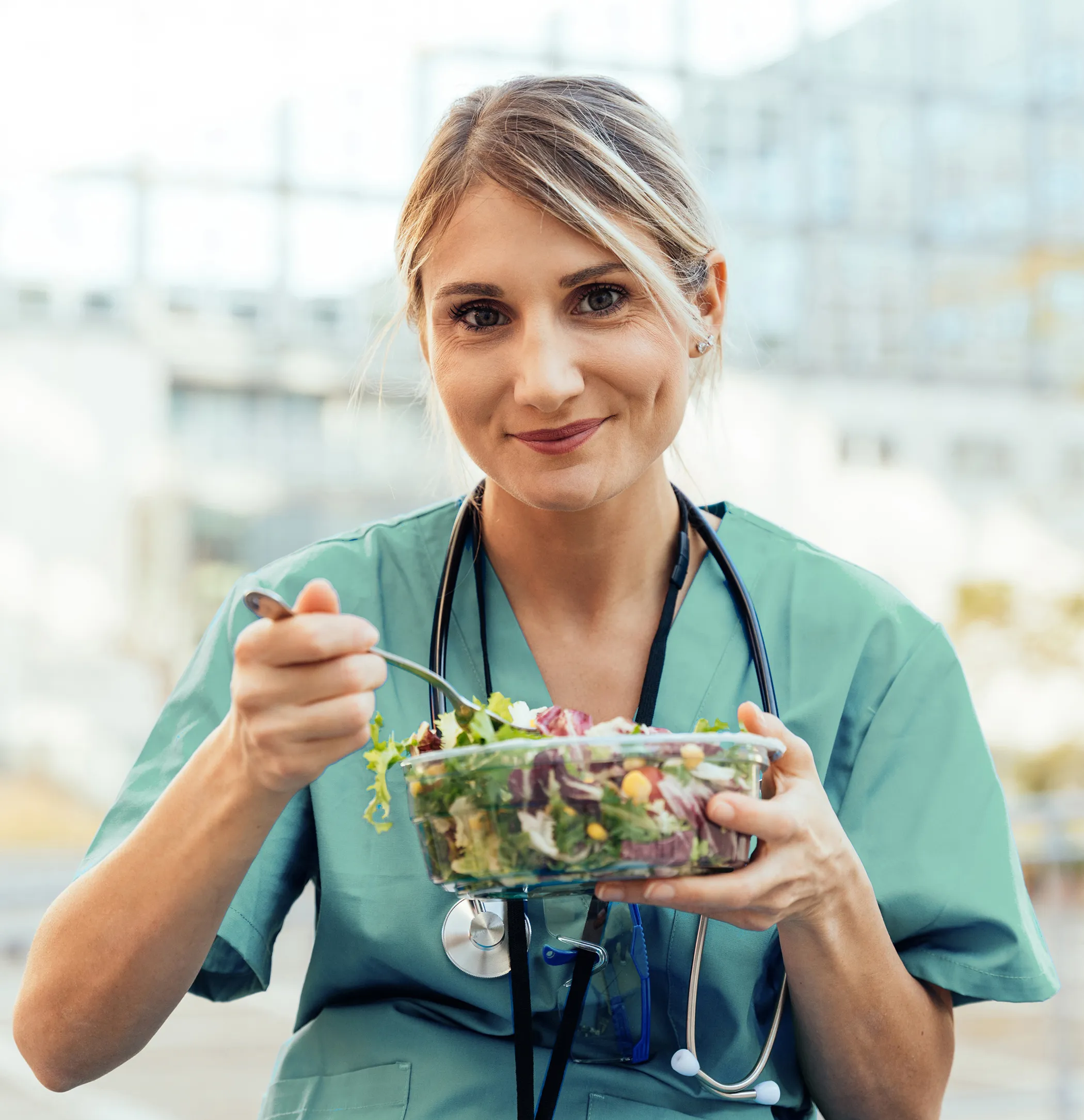 Une femme porte une blouse verte et elle a un stéthoscope autour de son cou. Elle mange une salade.