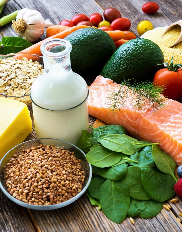 Une table sur laquelle il y a divers aliments, dont du poisson, du lait, des céréales, des épinards, des avocats, des carottes, des tomates, de l’ail et du beurre.