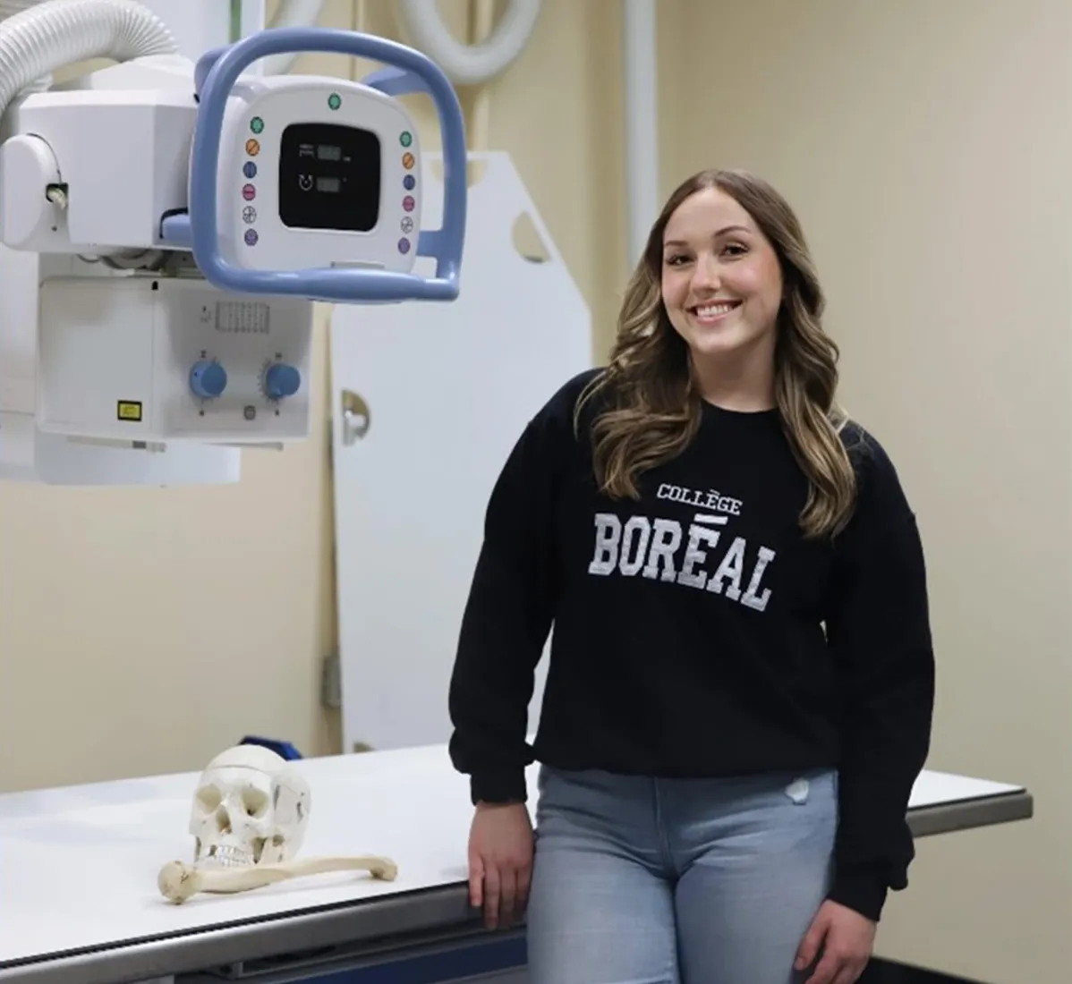 Emma Bissonnette est près d’un appareil de radiologie. Sur la table de radiologie, il y a un crâne.