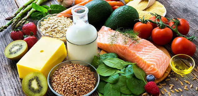 Une table sur laquelle il y a divers aliments, dont du poisson, du lait, des céréales, des épinards, des avocats, des carottes, des tomates, de l’ail et du beurre.