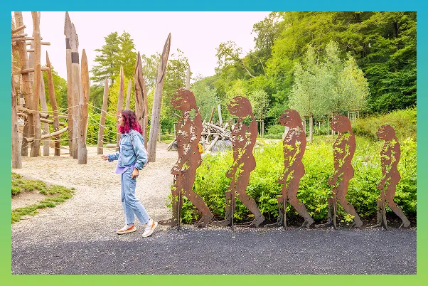 Une femme est dans un jardin. Elle pose devant des sculptures de bois représentant l’évolution de l’être humain.