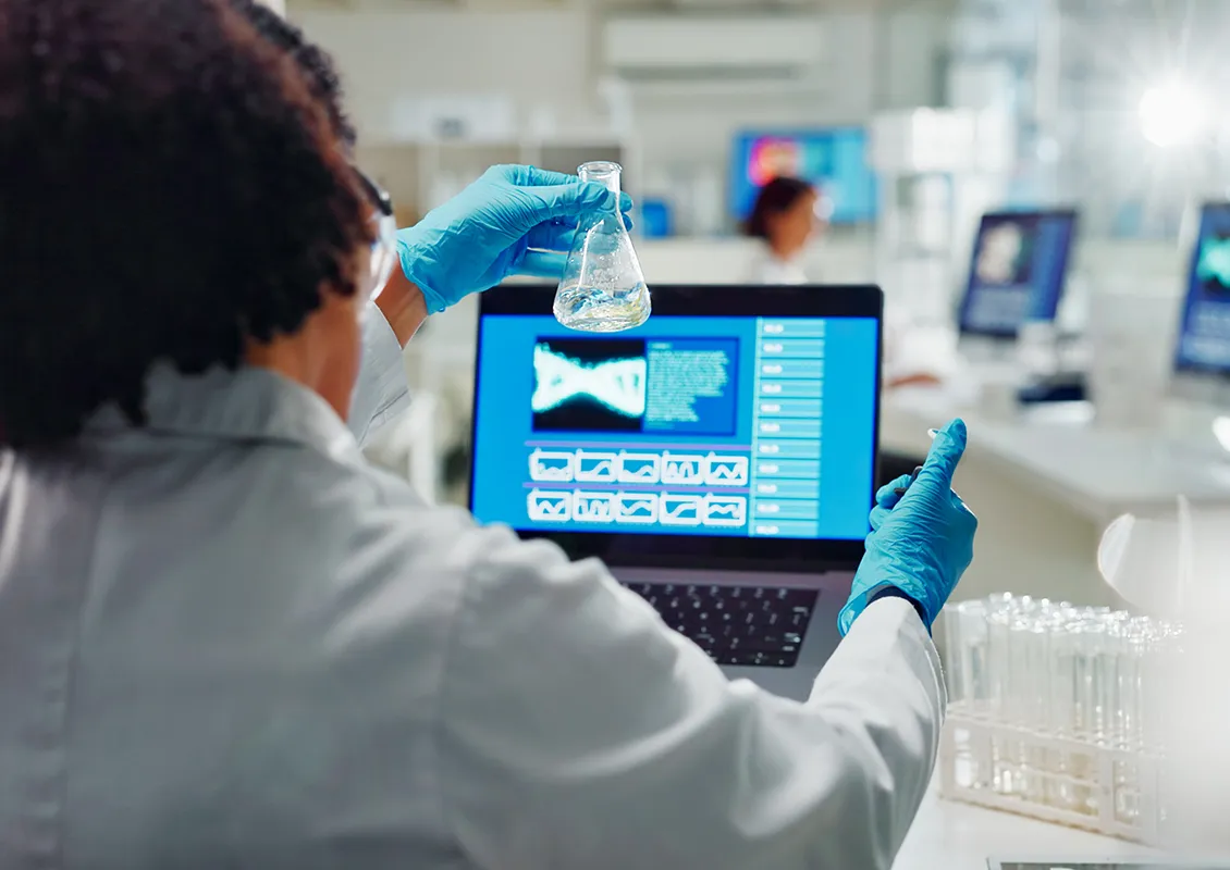 Une personne examine un flacon rempli de liquide tout en consultant des données sur un ordinateur portable.