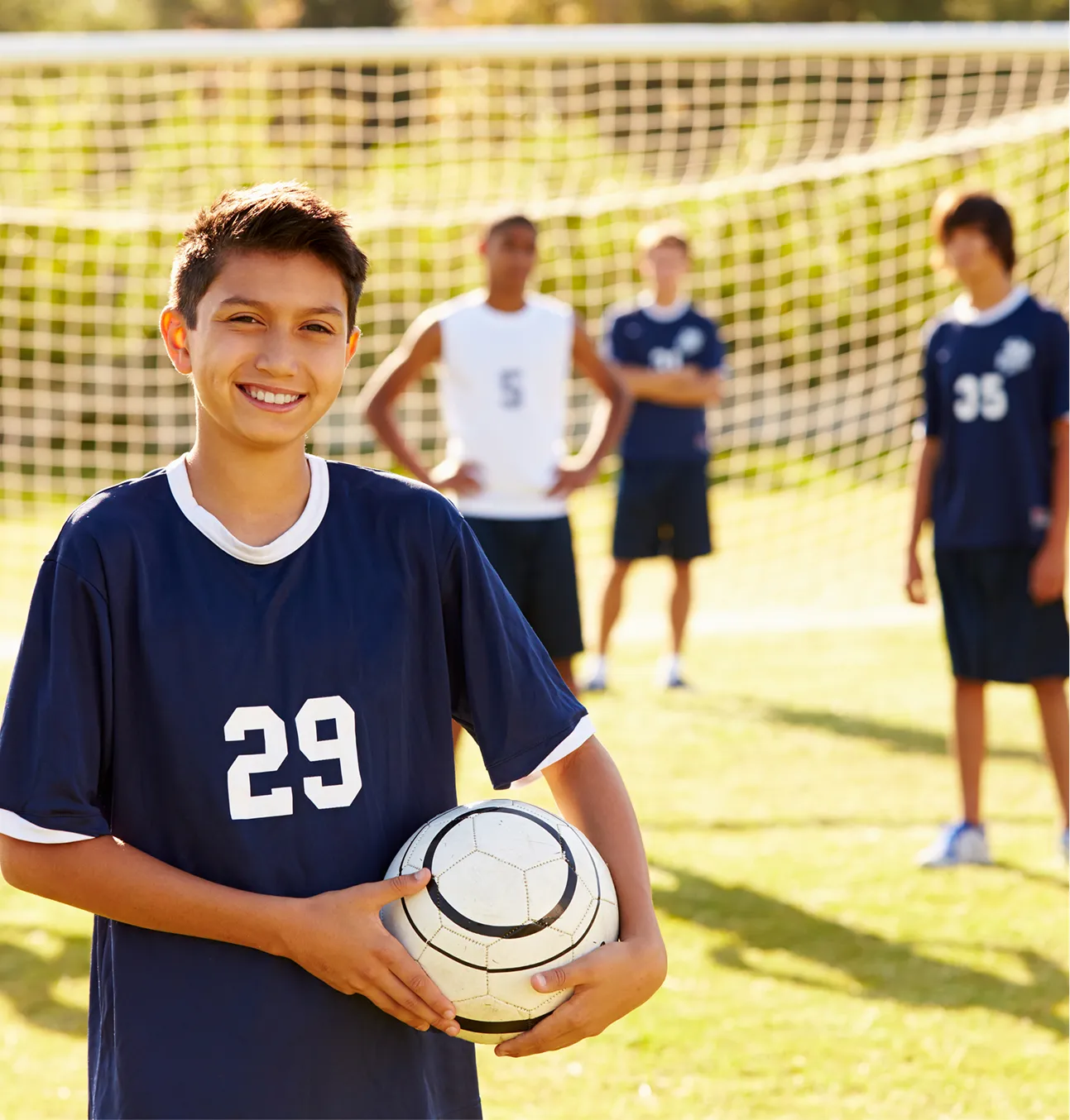 Un garçon tient un ballon entre ses mains. Derrière lui, il y a d’autres garçons qui l’attendent près d’un but de soccer.