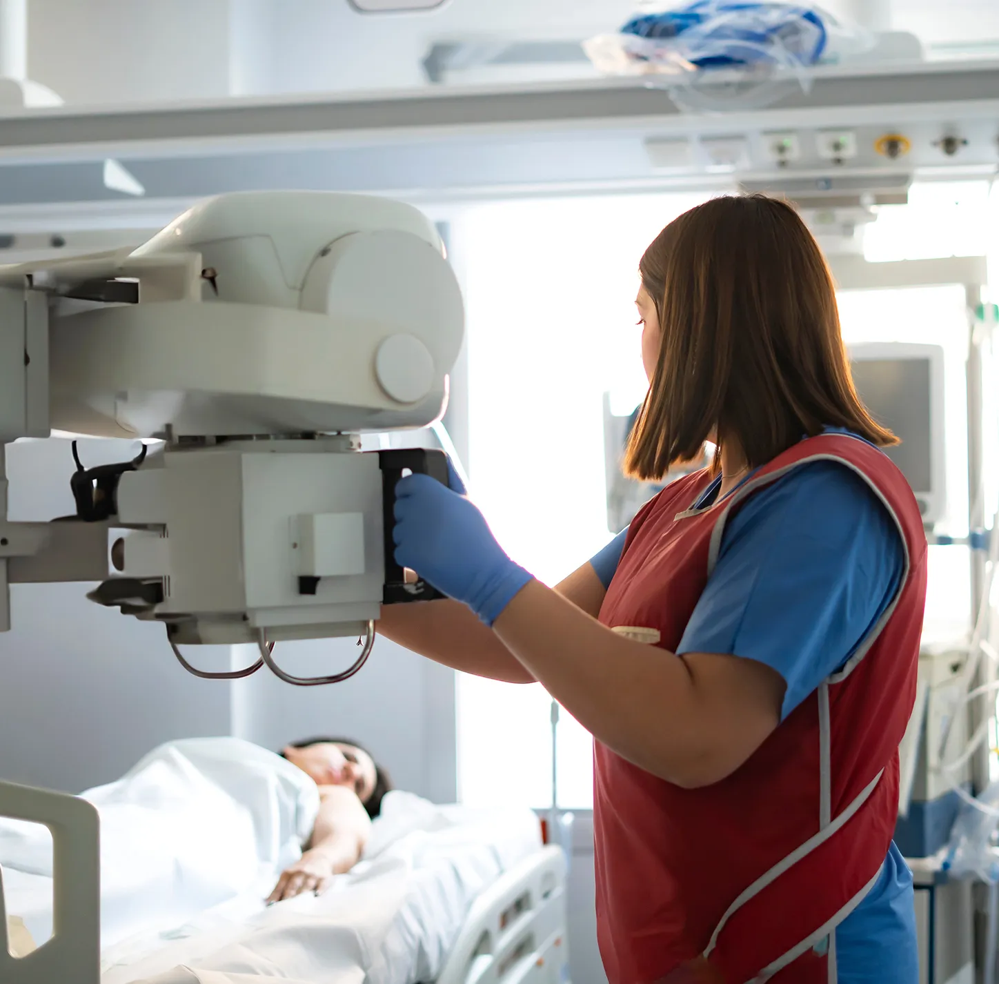 Une personne portant un tablier de plomb approche un appareil de radiologie vers une personne couchée dans un lit d’hôpital.