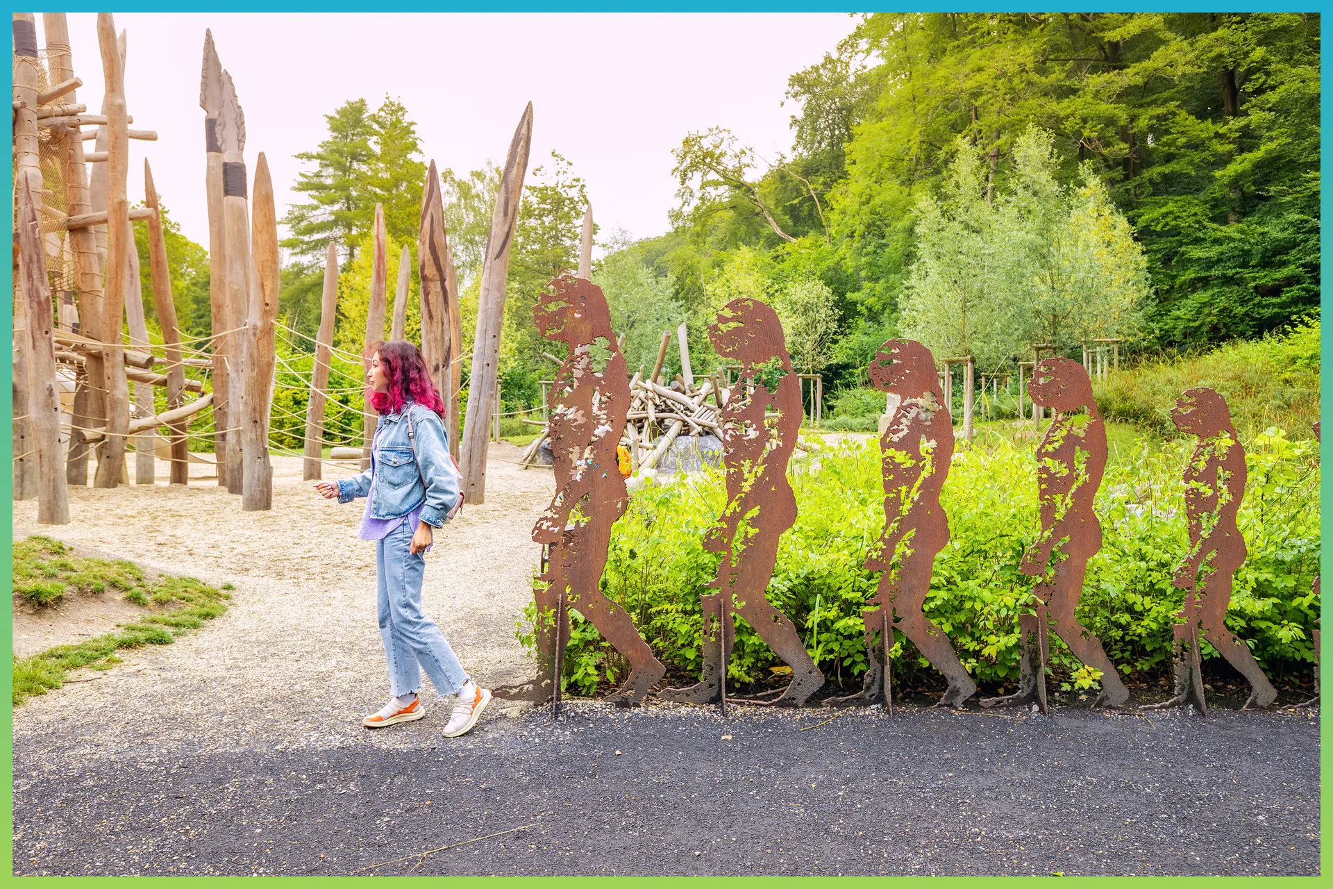 Une femme est dans un jardin. Elle pose devant des sculptures de bois représentant l’évolution de l’être humain.